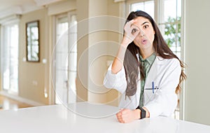 Young woman wearing medical coat at the clinic as therapist or doctor doing ok gesture shocked with surprised face, eye looking