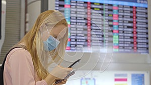 A young woman wearing a medical face mask in an airport stands in a hall in front of a flight schedule monitor with a