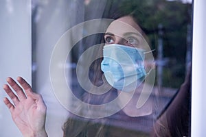 Young woman wearing a medical face mask for coronavirus and looking outside thought a Window. Covid-19