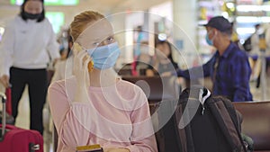 A young woman wearing a medical face mask sits on a chair in an airport and talks on a cellphone. The concept of the New