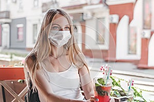 A young woman wearing a medical mask sits at a table in a roadside cafe alone and looks into the distance