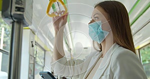 Young woman wearing medical mask to protect against coronavirus uses her smartphone while traveling on public transport