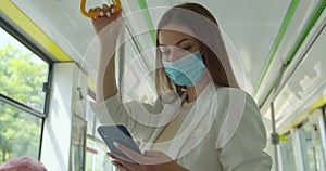 Young woman wearing medical mask to protect against coronavirus uses her smartphone while traveling on public transport