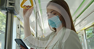 Young woman wearing medical mask to protect against coronavirus uses her smartphone while traveling on public transport