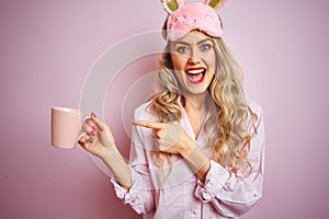 Young woman wearing pajama and mask drinking a cup of coffee over pink isolated background very happy pointing with hand and