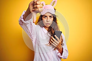 Young woman wearing pajama and mask using smartphone over yellow isolated background with angry face, negative sign showing