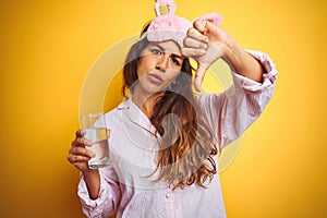 Young woman wearing pajama and sleep mask drinking water over yellow isolated background with angry face, negative sign showing