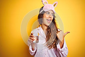 Young woman wearing pajama and sleep mask drinking water over yellow isolated background pointing and showing with thumb up to the