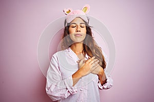 Young woman wearing pajama and sleep mask standing over pink isolated background smiling with hands on chest with closed eyes and