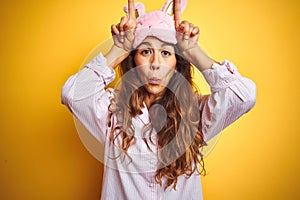 Young woman wearing pajama and sleep mask standing over yellow isolated background doing funny gesture with finger over head as