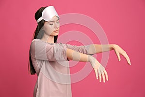 Young woman wearing pajamas and mask in sleepwalking state on pink background