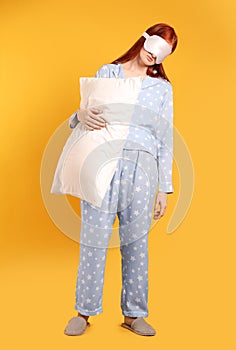 Young woman wearing pajamas and slippers with pillow in sleepwalking state on yellow background