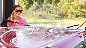 Young woman wearing pink t shirt and sunglasses cleaning her car in self serve carwash, blurred sun lit trees background