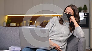 A young woman wearing protective face mask sits on the couch with a laptop