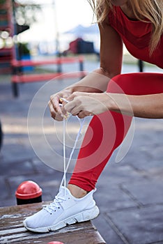 Young woman, wearing red fitness outfit, tying shoe laces on white sneakers. Process of fastening sport shoes. Close-up shot of