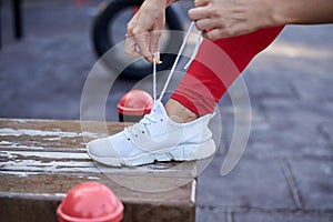 Young woman, wearing red fitness outfit, tying shoe laces on white sneakers. Process of fastening sport shoes. Close-up shot of