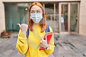 Young woman wearing safety mask and student backpack holding books pointing thumb up to the side smiling happy with open mouth