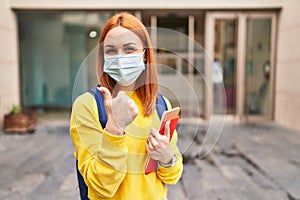 Young woman wearing safety mask and student backpack holding books smiling happy and positive, thumb up doing excellent and