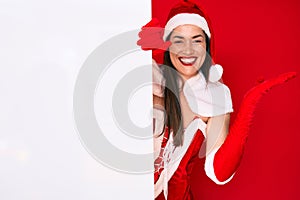 Young woman wearing santa claus costume holding blank empty banner celebrating victory with happy smile and winner expression with