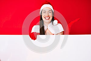 Young woman wearing santa claus costume holding blank empty banner celebrating victory with happy smile and winner expression with