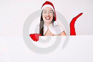 Young woman wearing santa claus costume holding blank empty banner celebrating victory with happy smile and winner expression with