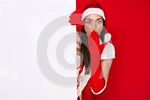Young woman wearing santa claus costume holding blank empty banner covering mouth with hand, shocked and afraid for mistake