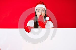 Young woman wearing santa claus costume holding blank empty banner covering mouth with hand, shocked and afraid for mistake