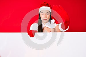 Young woman wearing santa claus costume holding blank empty banner with open hand doing stop sign with serious and confident