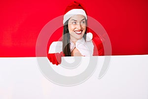 Young woman wearing santa claus costume holding blank empty banner pointing thumb up to the side smiling happy with open mouth
