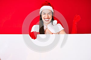 Young woman wearing santa claus costume holding blank empty banner screaming proud, celebrating victory and success very excited