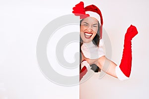 Young woman wearing santa claus costume holding blank empty banner screaming proud, celebrating victory and success very excited