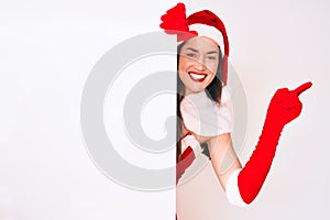 Young woman wearing santa claus costume holding blank empty banner smiling happy pointing with hand and finger to the side