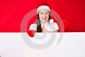 Young woman wearing santa claus costume holding blank empty banner smiling happy pointing with hand and finger to the side