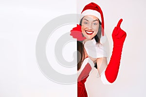 Young woman wearing santa claus costume holding blank empty banner surprised with an idea or question pointing finger with happy