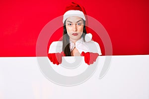 Young woman wearing santa claus costume holding blank empty banner thinking attitude and sober expression looking self confident