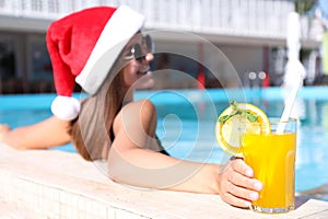 Young woman wearing Santa Claus hat with refreshing drink in swimming pool, focus on hand. Christmas vacation