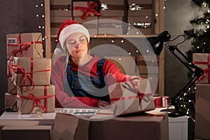 A young woman wearing a Santa Claus hat works in a warehouse of boxes with gifts and orders from an online store for