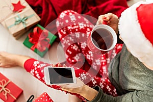 Young woman wearing santa hat and xmas pajamas sitting on the floor amongst wrapped christmas presents, shopping on her phone.