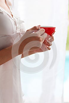 A young woman wearing a sexy white see through nightgown stands by the window with a red cup of coffee in the morning to drink