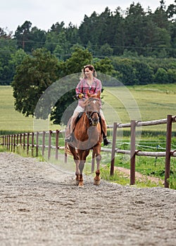 Young woman wearing shirt riding brown horse in sand paddock by wooden fence, hair moving in air because of speed