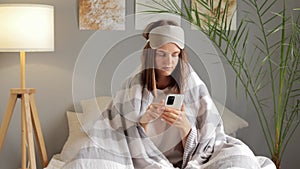 Young woman wearing sleep eye mask wrapped in duvet blanket holding mobile cell phone at home scrolling online reading news after