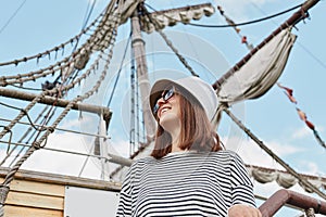 Young woman wearing striped shirt panama sunglasses standing on ship with sails and sky on background looking away enjoying