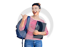 Young woman wearing student backpack holding binder pointing thumb up to the side smiling happy with open mouth