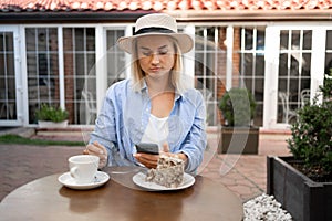 Young woman wearing summer hat typing text message on smart phone in a cafe. Girl with a coffee and dessert using mobile