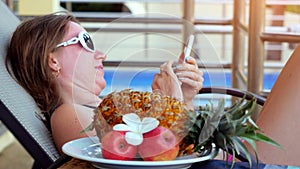 Young woman wearing sunglasses lying on a sunbed uses smartphone sending a message next to the swimming pool on summer