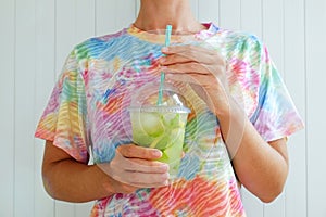 Young woman wearing a tie dye t-shirt holding a glass of iced lemonade. Female with refreshing non alcoholic mojito drink with