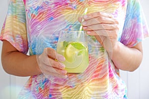 Young woman wearing a tie dye t-shirt holding a glass of iced lemonade. Female with refreshing non alcoholic mojito drink with