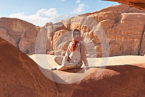 Young woman wearing warm jacket and scarf around head smiling, sitting at naturally formed rock window in wadi rum desert on sunny