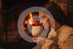 Young woman wearing white woolen knitted sweater enjoying hot tea near fireplace in a cozy living room