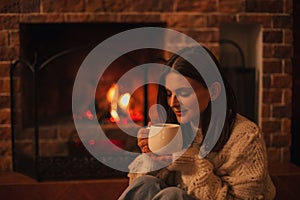 Young woman wearing white woolen knitted sweater enjoying hot tea near fireplace in a cozy living room